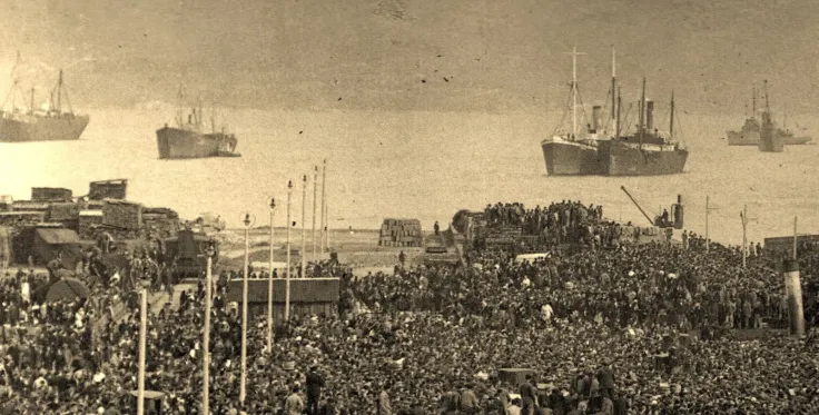 A dense crowd on a quayside with steamships lying at anchor beyond