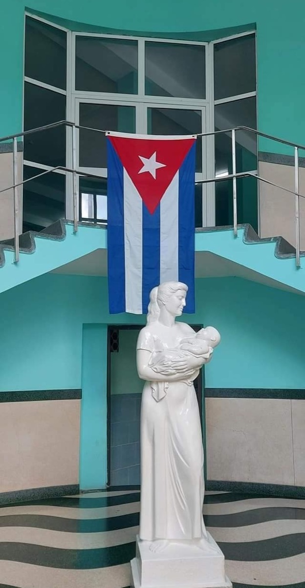 A statue of a mother holding an infant beneath a Cuban flag, in the stairwell of the Hijas de Galicia hospital